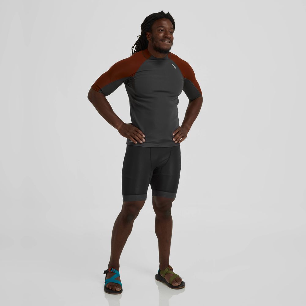 A person with long hair wears a gray and red NRS Hydroskin 0.5mm Short Sleeve Shirt - Men's, black shorts, and sandals. They stand with hands on hips, smiling against a plain white background.