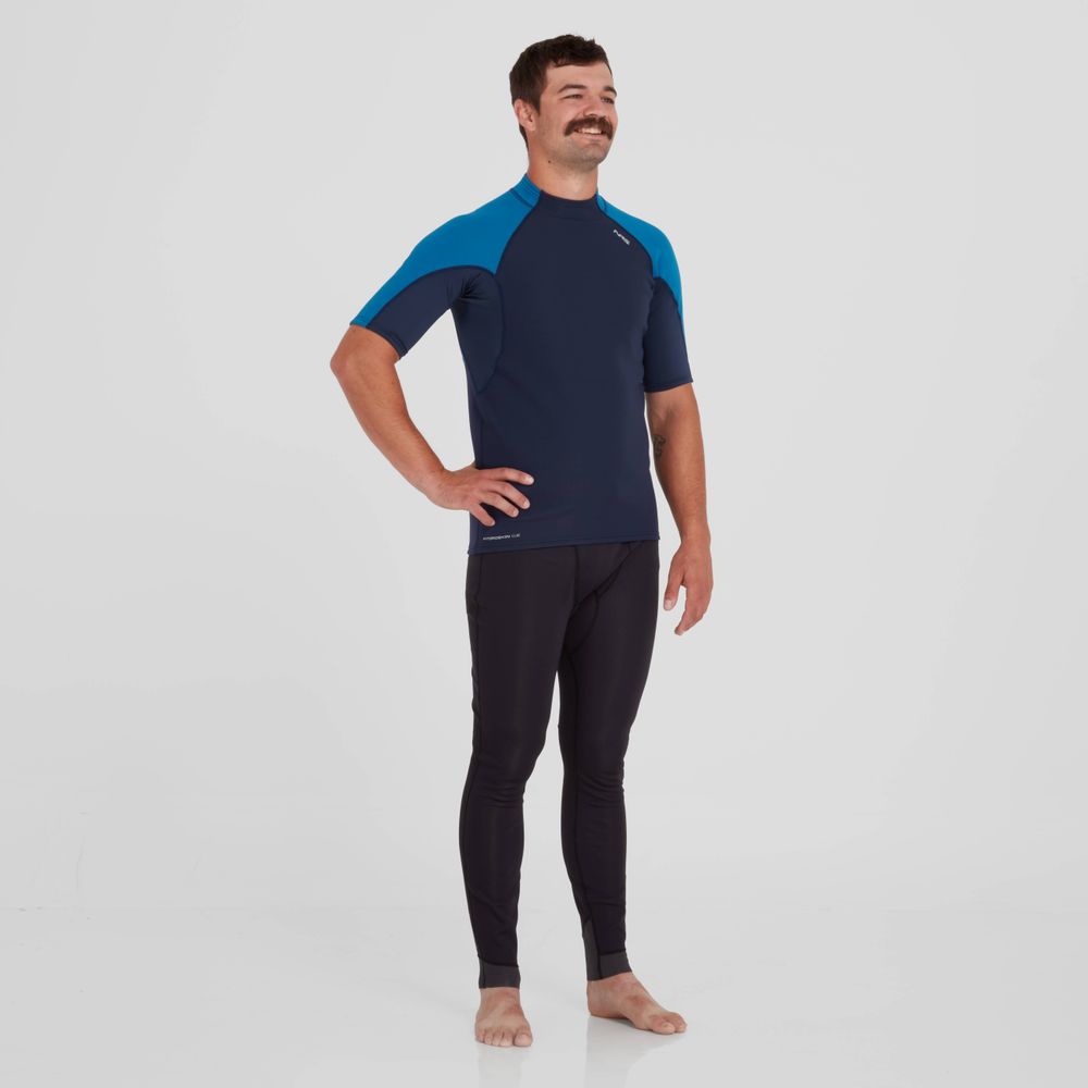 A man stands barefoot, wearing a two-tone blue and dark athletic outfit with a short-sleeved NRS Hydroskin 0.5mm Short Sleeve Shirt - Men's and leggings, posing against a white background.