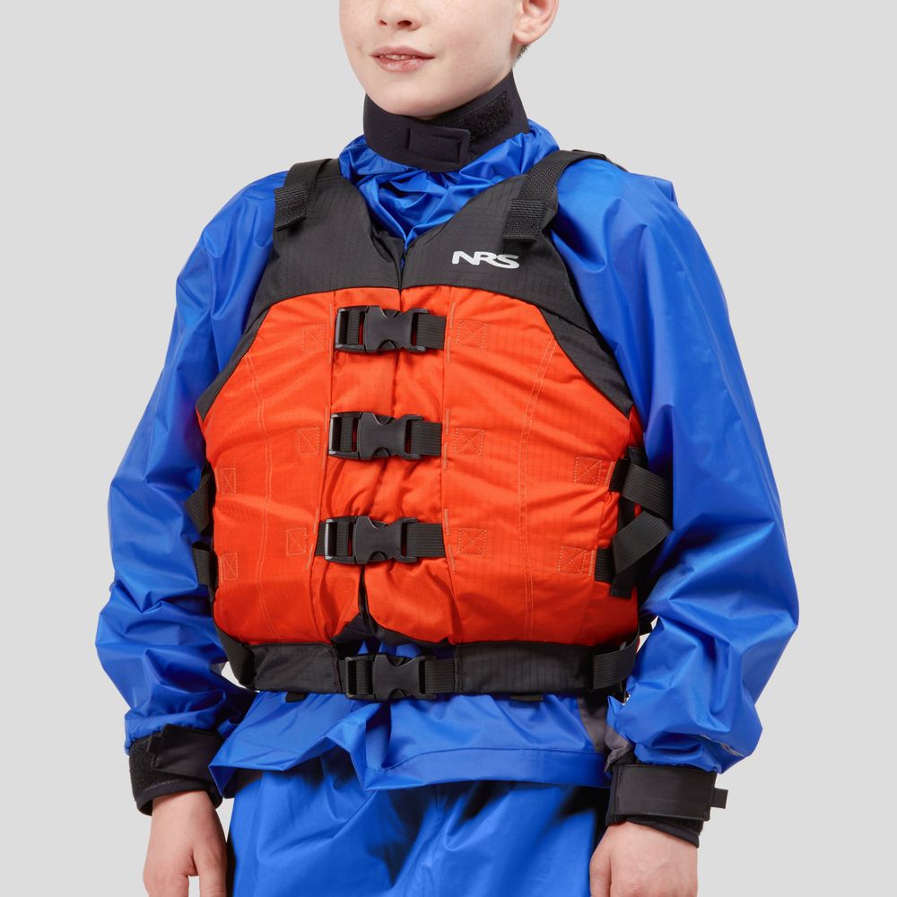 A person stands against a gray background, wearing a blue dry suit paired with the NRS Big Water V Youth PFD in orange, featuring black straps.