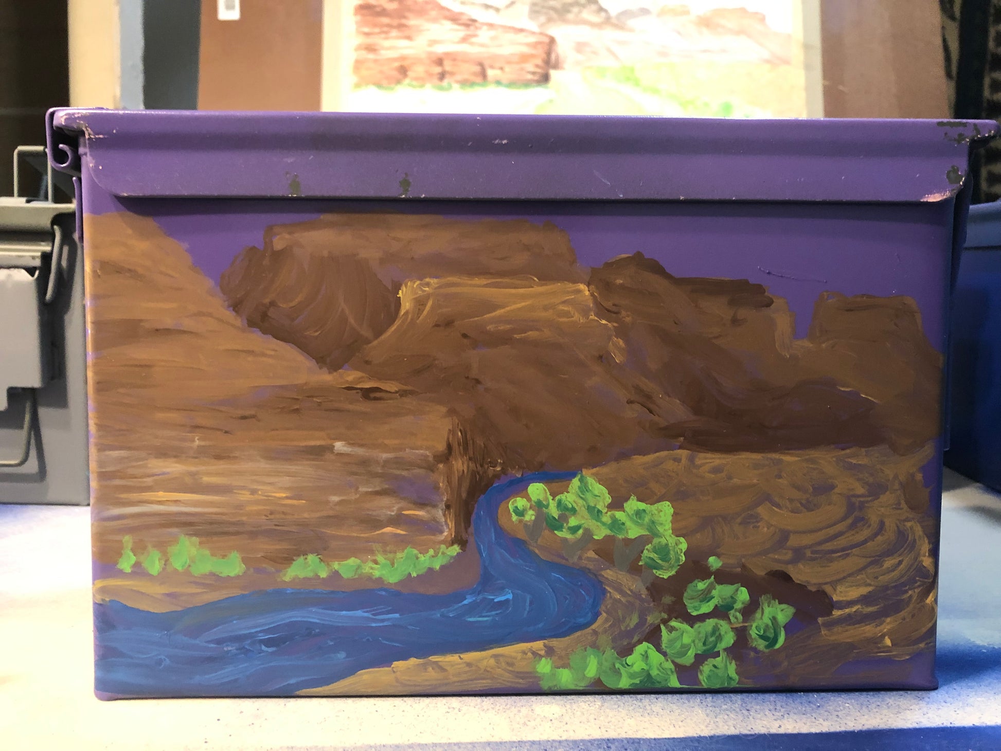 The Paint and Sip- Ammo Can! by 4CRS Paddle School features a purple box with a painted landscape by Aaron Schenk, depicting brown mountains, a winding blue river, and green trees.