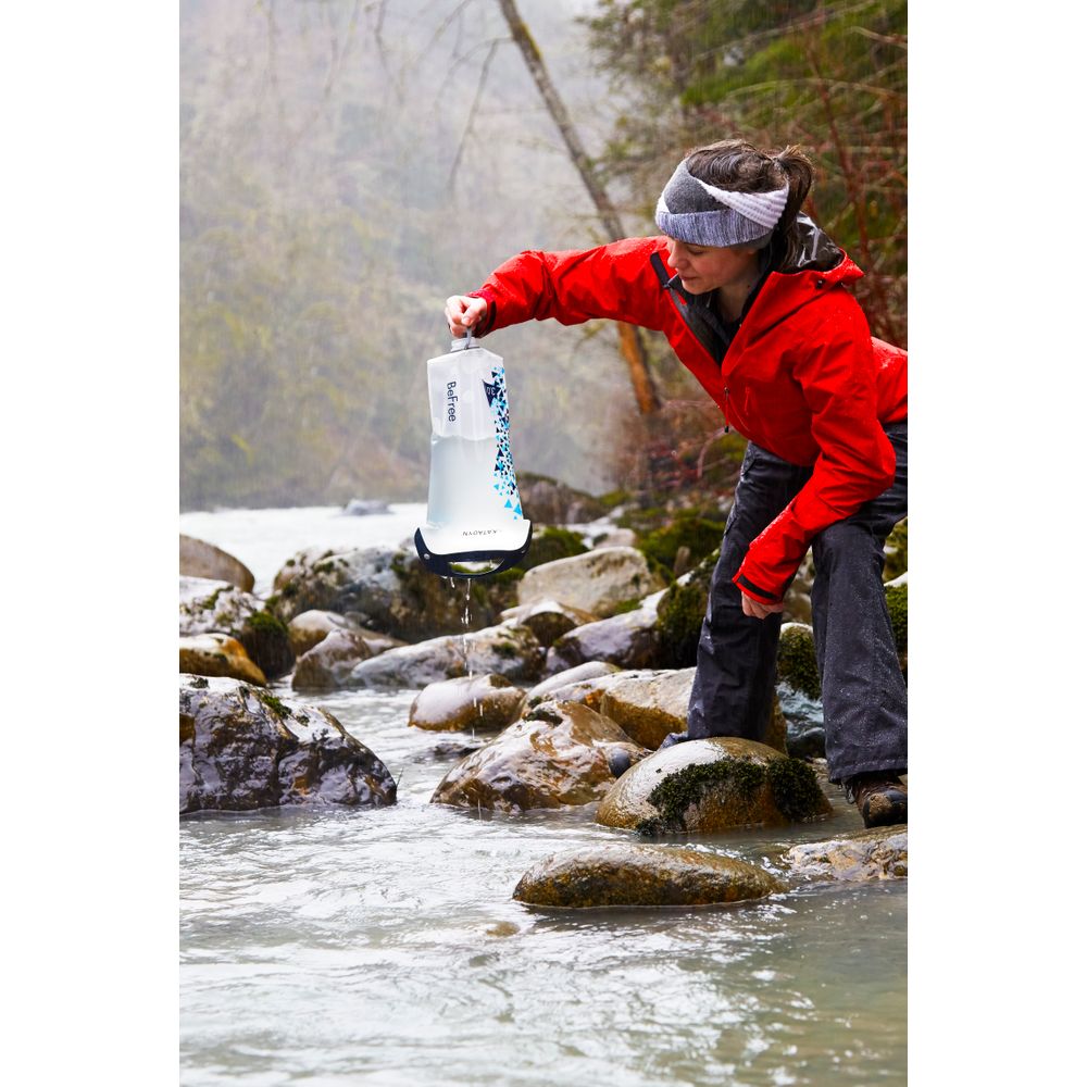 A person in a red jacket collects water from a rocky stream using the NRS Base Camp Pro Water Filter, featuring a robust pleated glass-fiber filter for efficient purification.