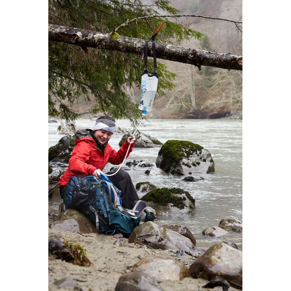 A person in a red jacket sits by a river with an NRS Base Camp Pro Water Filter, efficiently purifying water with its advanced pleated glass-fiber filter, as it hangs among moss-covered rocks and trees, filling their backpack.