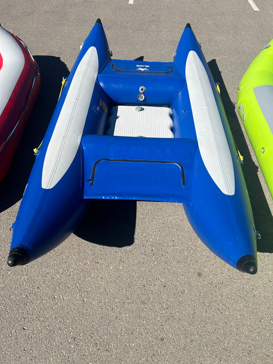 Blue inflatable boat with gray seats and a storage compartment, an unmistakable member of the rental fleet boat collection, positioned between two other inflatable boats on a concrete surface. This Used Demo Slice from NRS boasts just 1 season of use.