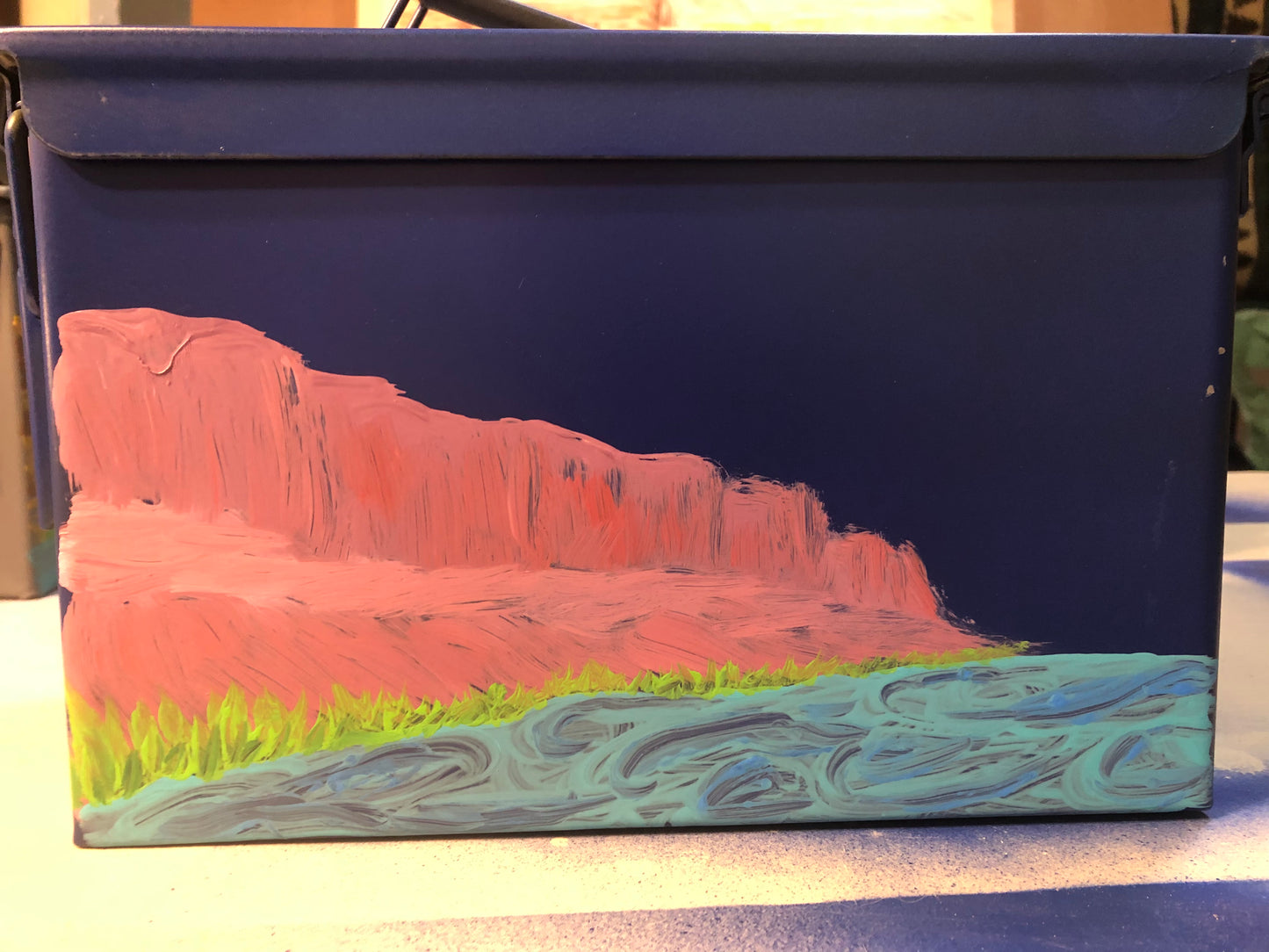 The Paint and Sip- Ammo Can! by 4CRS Paddle School features a vibrant acrylic painting of red rock formations, lush green grass, and a flowing blue river under a deep blue sky on a metal surface crafted in a guided class.