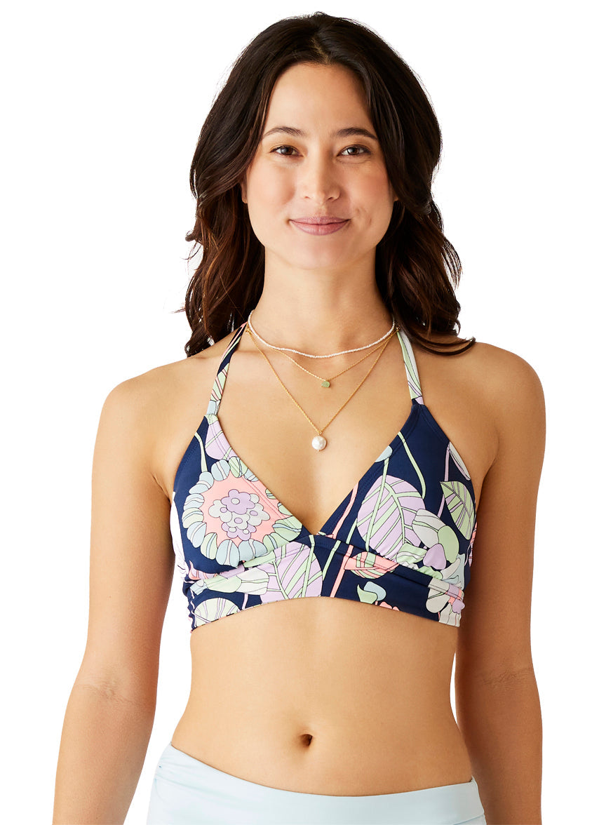 Confidently posing against a white background, the woman wears a Carve Dahlia Top with a flattering fit, her floral sports bra complementing her layered necklaces. She smiles brightly, embodying style and elegance in her active swimwear look.