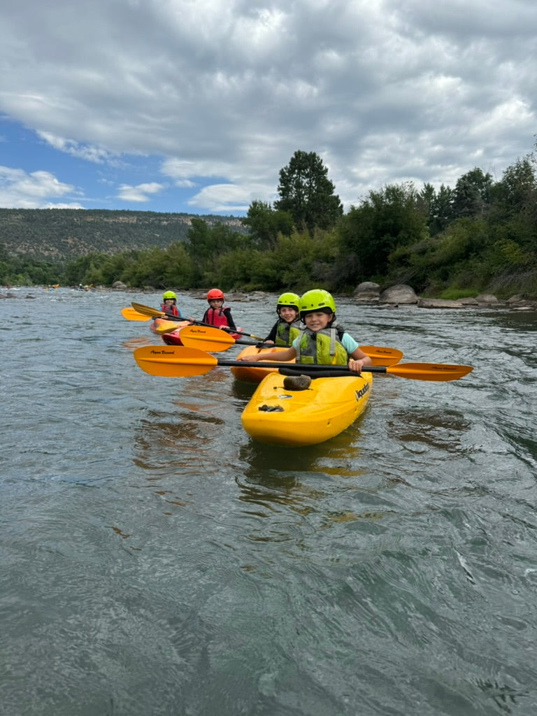 A group of four children in yellow kayaks, possibly participants from a 4Corners Riversports Kids Paddle School class, drift along a river framed by trees and hills under an overcast sky.