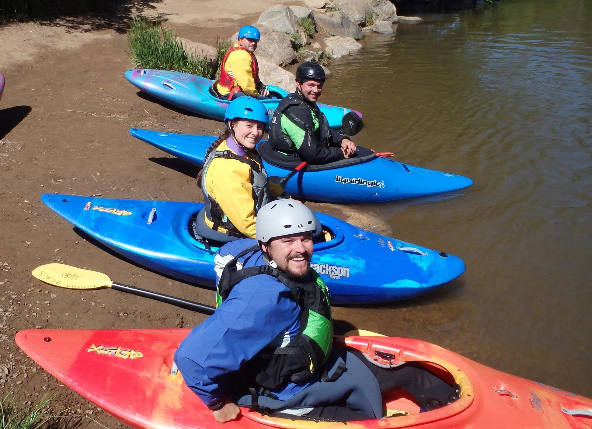 Four kayakers in vibrant gear sit in kayaks by the water's edge, smiling at the camera and eager for their next adventure with the Adult Paddle School Class Holiday Gift Certificate from 4Corners Riversports.