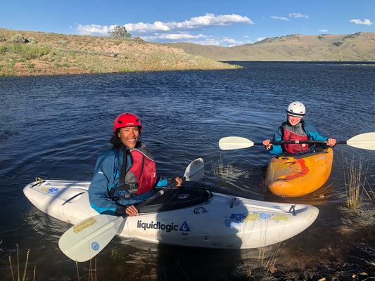 Two adult kayakers, equipped with life jackets and helmets from 4CRS Paddle School's Adult Intro to Kayaking Class, are mastering paddle strokes while sitting in their kayaks by the lake. Encircled by grassy hills under a clear sky, they're embracing the basics of boating in this serene setting.