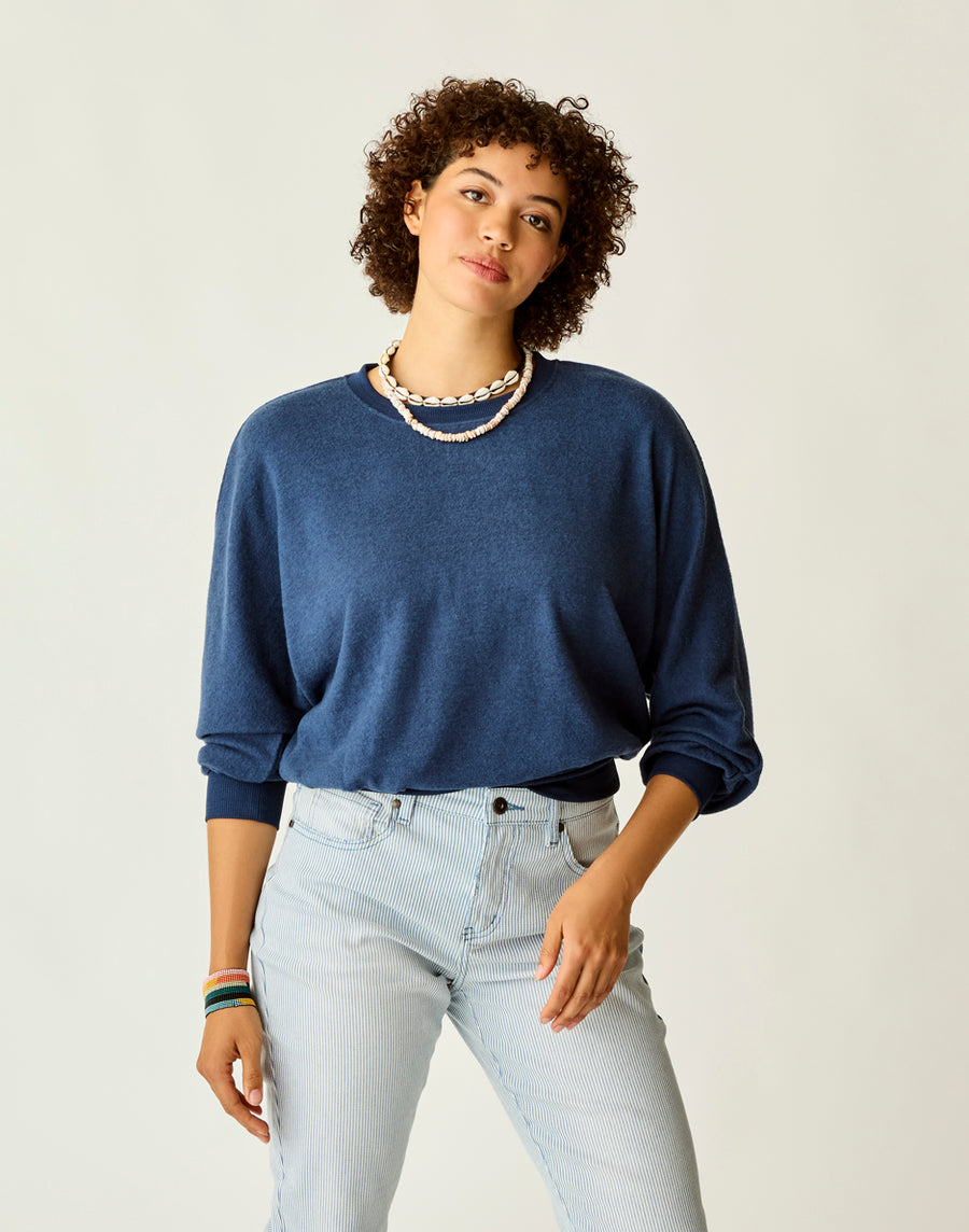 An individual with short curly hair is wearing a blue "Anneke Supersoft Dolman" sweater by Carve, paired with light blue jeans and a seashell necklace, standing against a plain background.