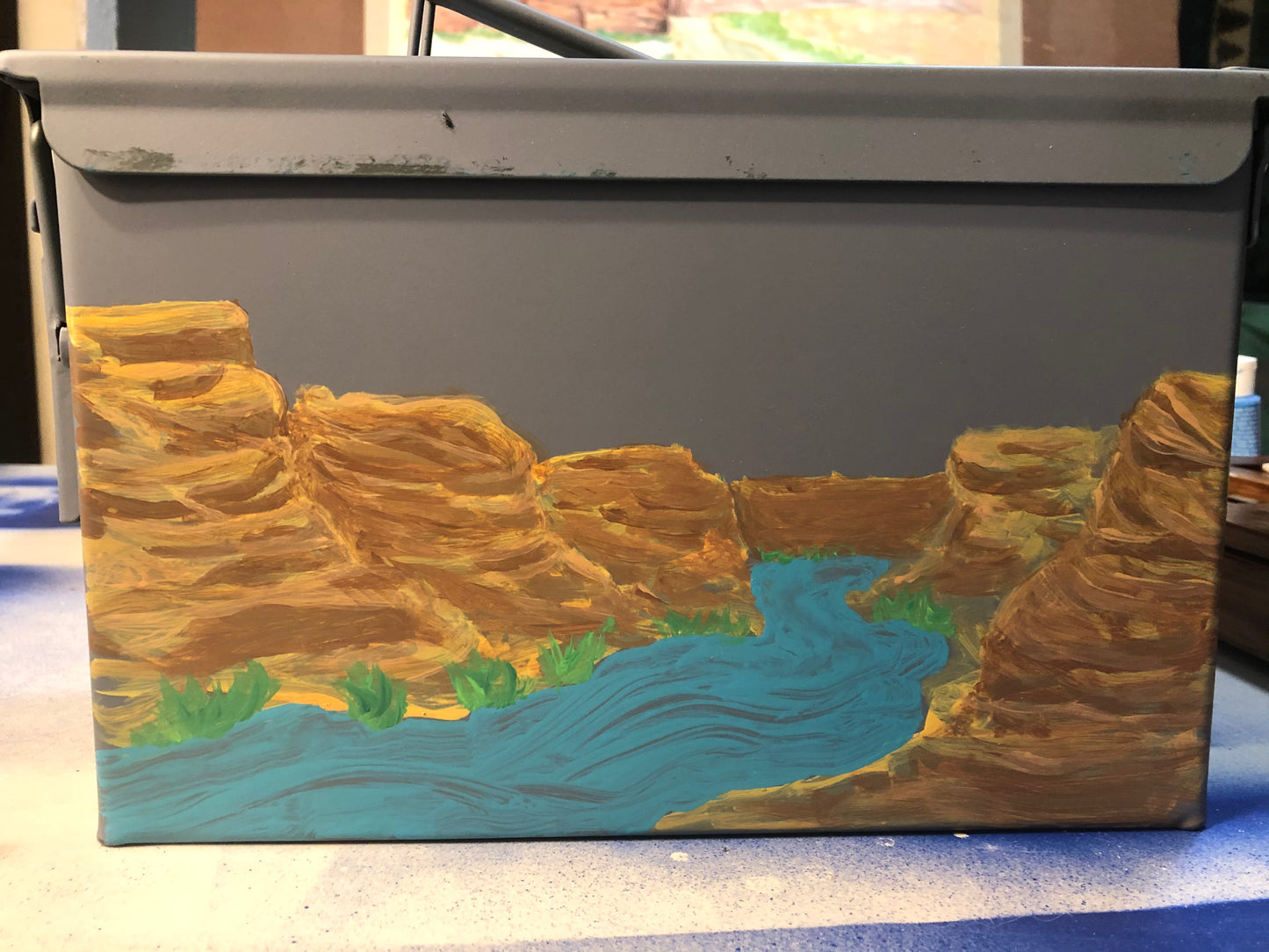 The Paint and Sip- Ammo Can by 4CRS Paddle School is a gray metal box featuring a hand-painted river in rocky canyons, created in a guided acrylic class by local artist Aaron Schenk.