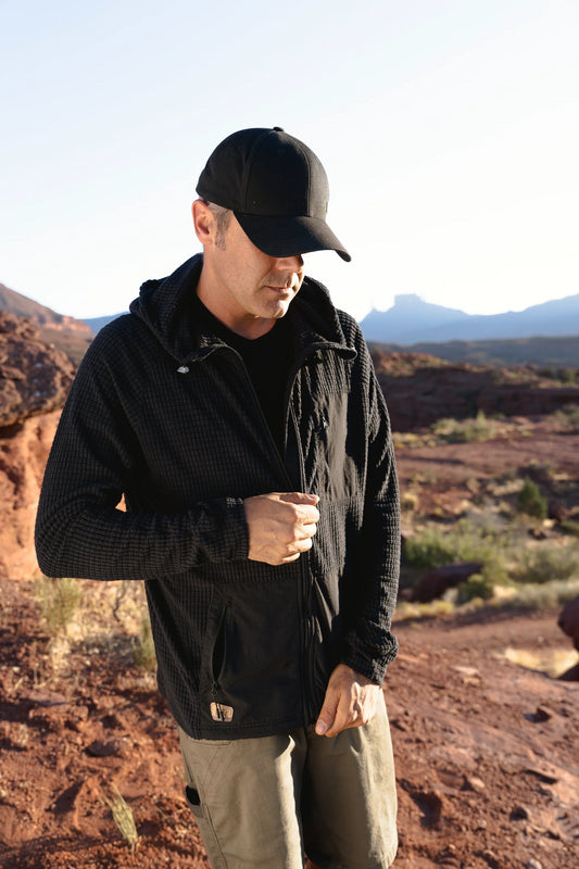 A person wearing a Castleton Grid Hoodie M's by Desert Wild zips up their jacket in a rocky desert landscape under a bright sky.