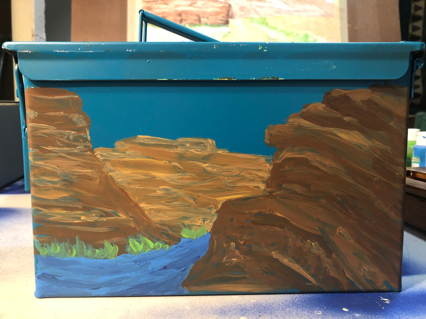 The Paint and Sip- Ammo Can by 4CRS Paddle School showcases a captivating acrylic river scene between brown cliffs on a blue metal toolbox, evoking the charm of a guided painting class.