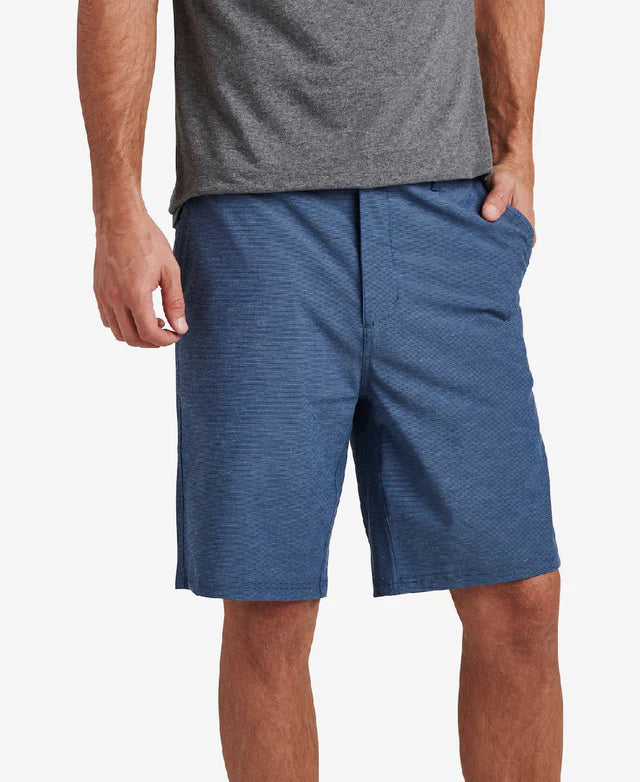 A person is standing against a plain background, casually wearing a gray t-shirt and REEF's Dante Short, which features a 20-inch outseam in blue made from a cotton/poly/spandex blend, with one hand in their pocket.