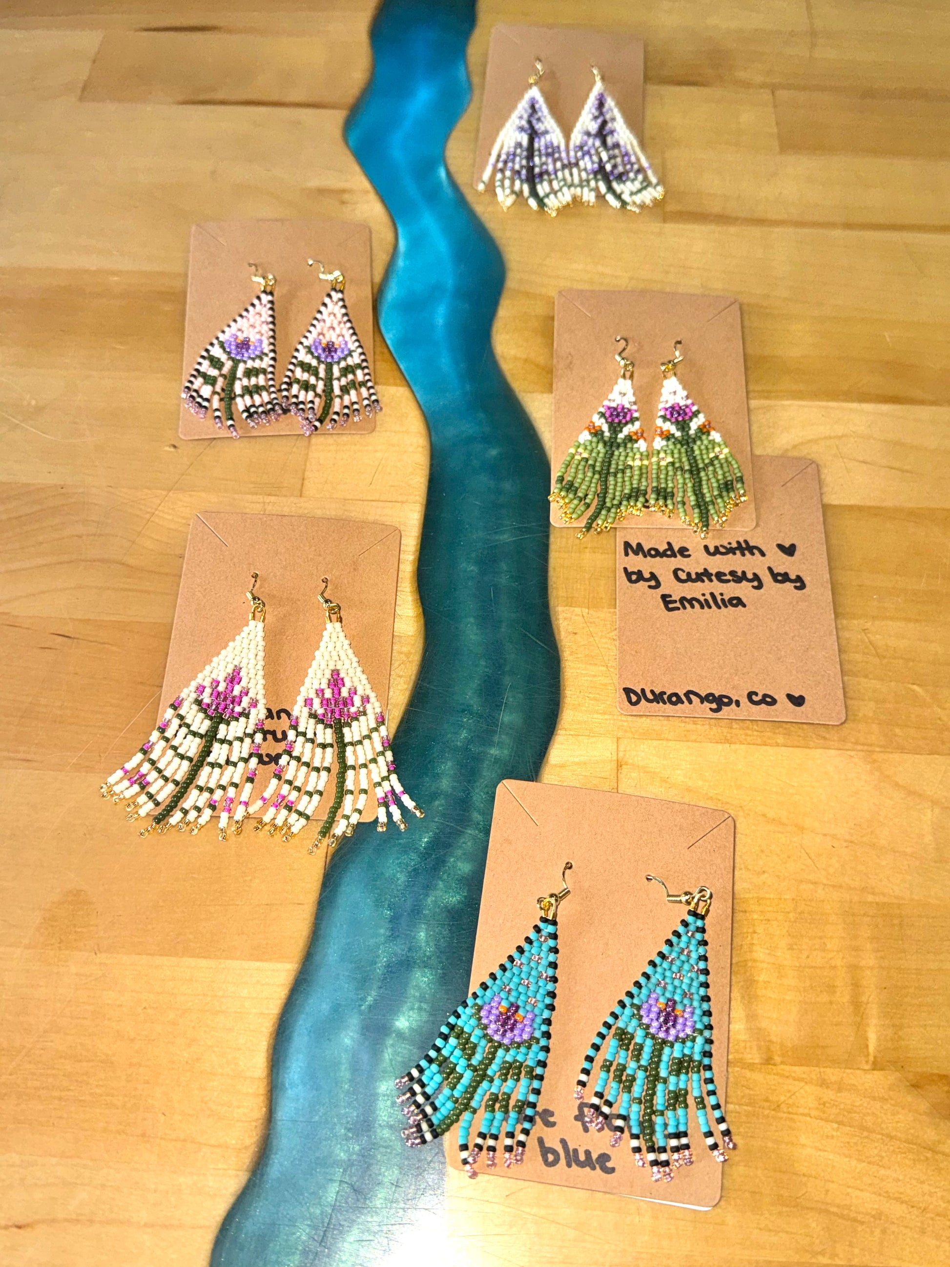 Five pairs of Cutesy By Emilia Earrings from 4CRS, featuring colorful beads on brown cards, are elegantly displayed on wood with a blue accent. Each piece showcases intricate artistry inspired by desert flowers, adding nature's beauty to your collection.