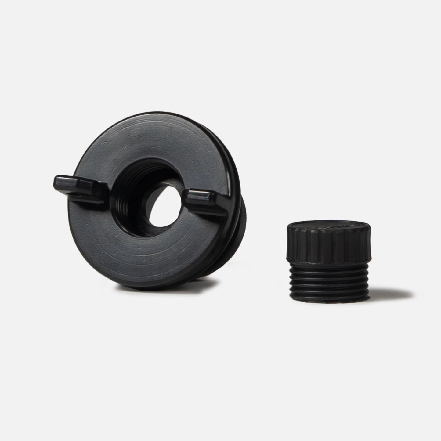 A sleek black circular cooler drain plug by Canyon Coolers, with two prongs, sits next to a small cylindrical cap on a plain white background, reminiscent of high-flow systems.