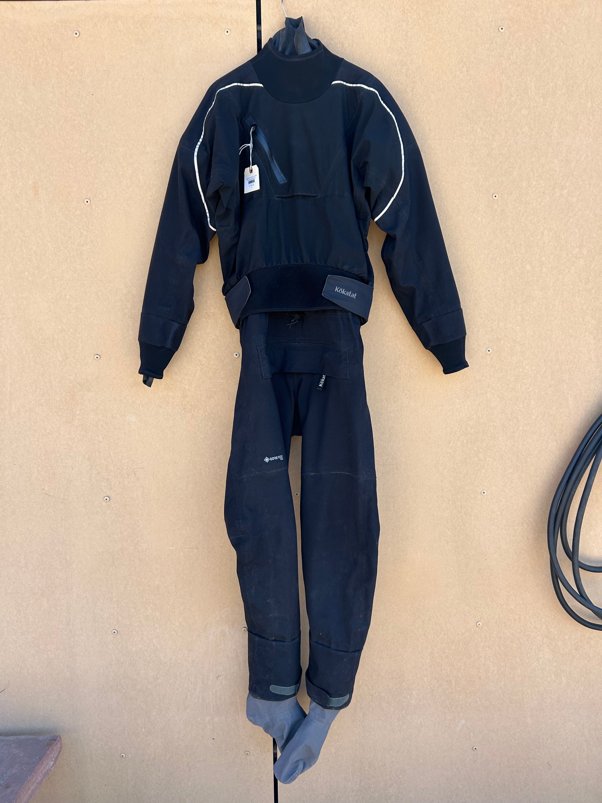 A Demo Drysuit- Kokatat Retro Icon Men's LG in black, featuring attached gray socks, hangs on a tan wall with a visible sales tag on the collar. A black hose is coiled to the right.