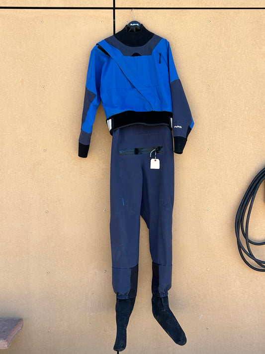 A blue and black Kokatat Demo Drysuit - NRS Axiom Men's XL hangs on a beige wall, with a hose coiled to the right side.