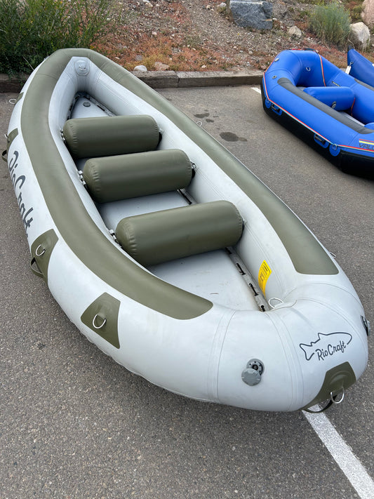 A gray inflatable raft with green padded seats, labeled "Consignment Teton 13.5'," from the brand Rio Craft, is parked on a paved surface near another blue raft.