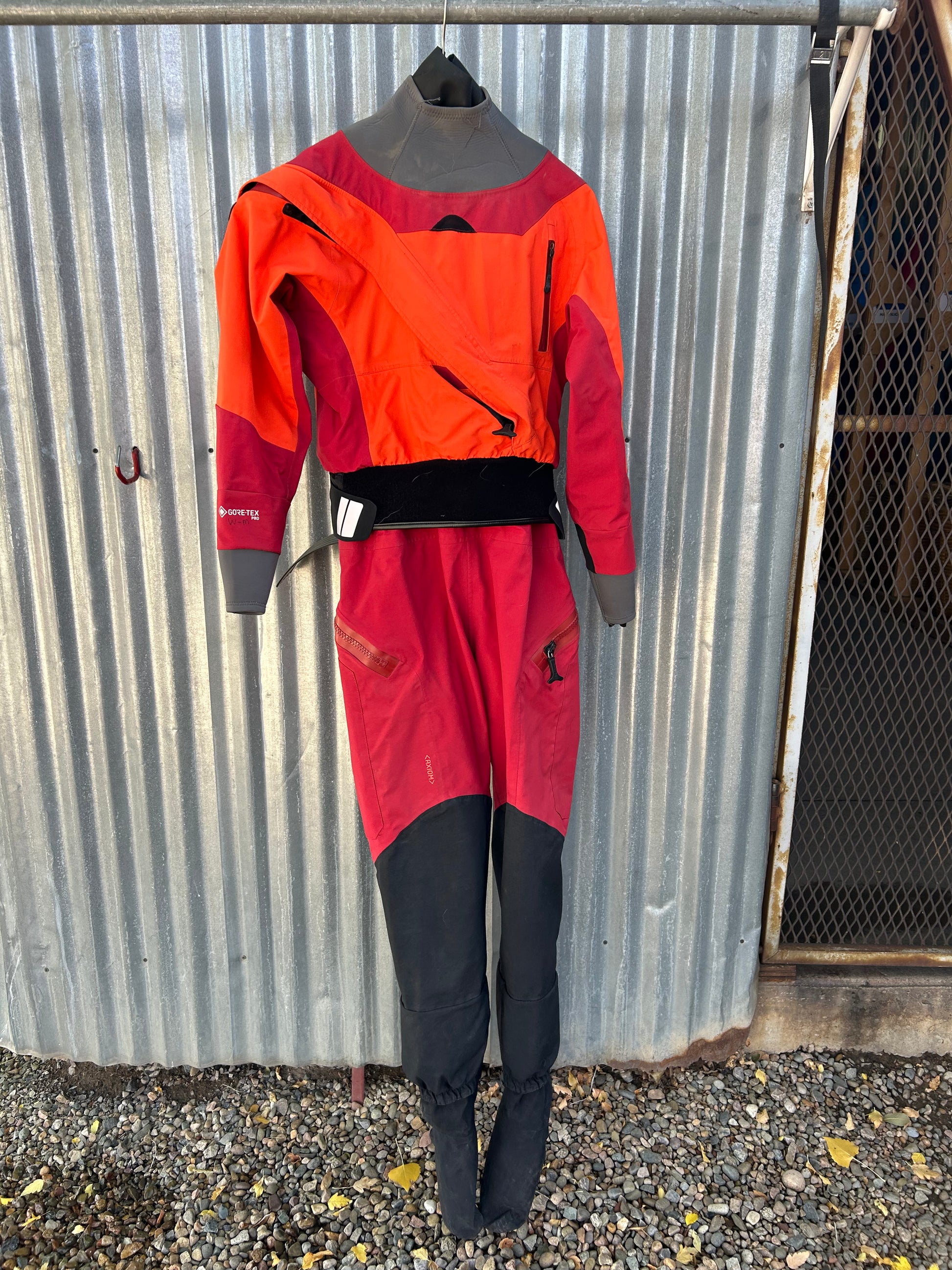 A Demo Drysuit - NRS Axiom W's MD, featuring orange and red colors with black accents, is hanging on a metal rod against a corrugated metal wall.