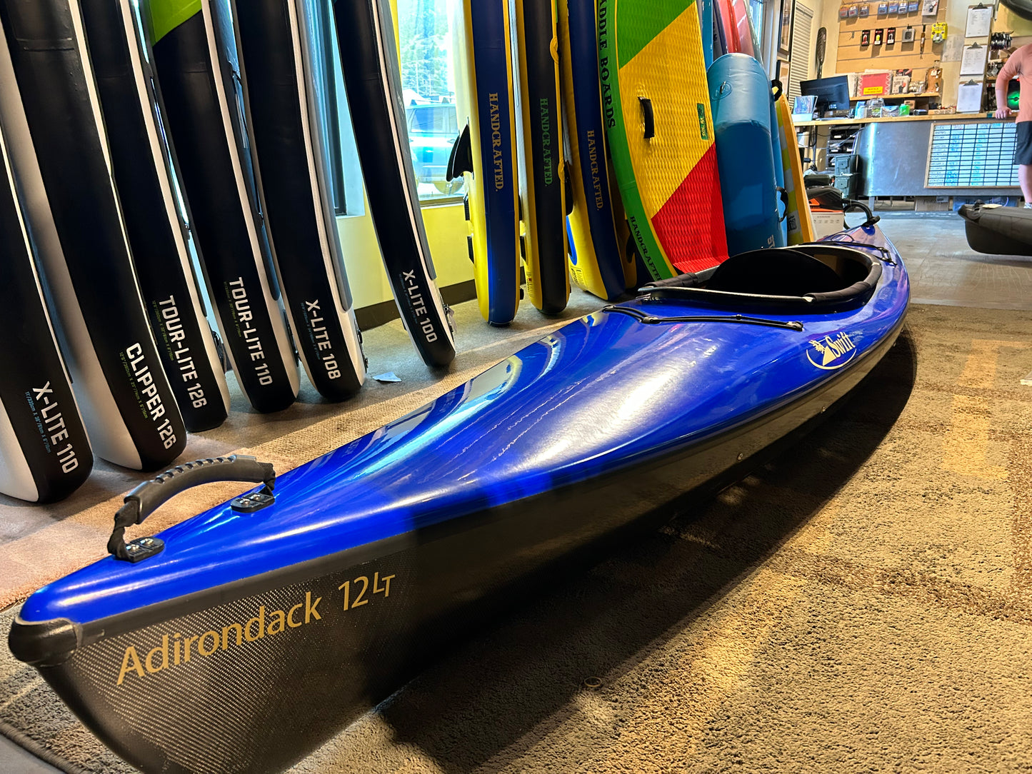 A blue 4CRS Consignment Swift Adirondack 12 LT kayak with a narrower cockpit is displayed on the floor, surrounded by various upright paddleboards and equipment in a store setting.