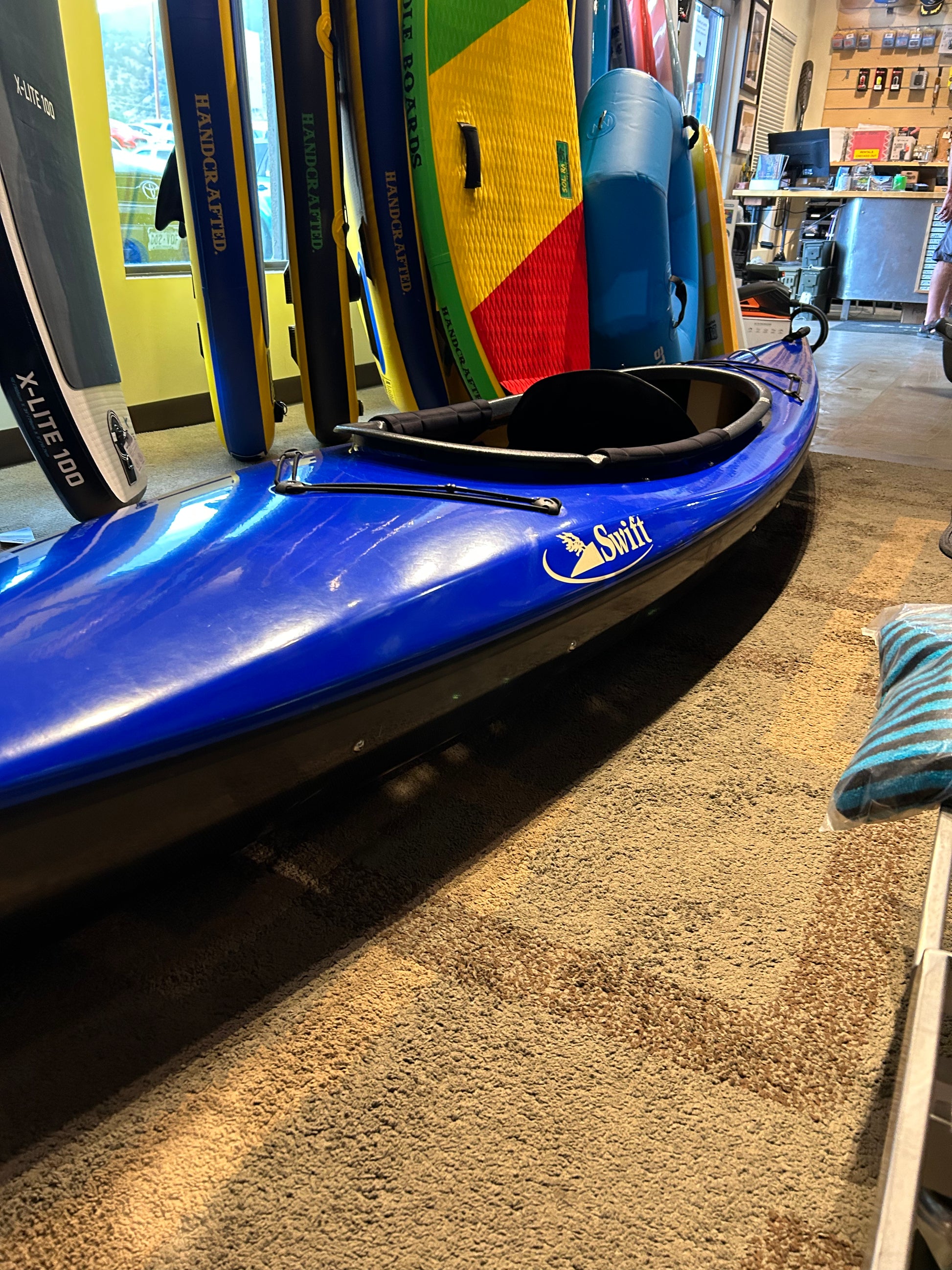 A blue kayak labeled "Consignment Swift Adirondack 12 LT" by 4CRS is on display indoors, featuring a narrower cockpit and surrounded by various colored paddleboards. Neon yellow walls are visible in the background.