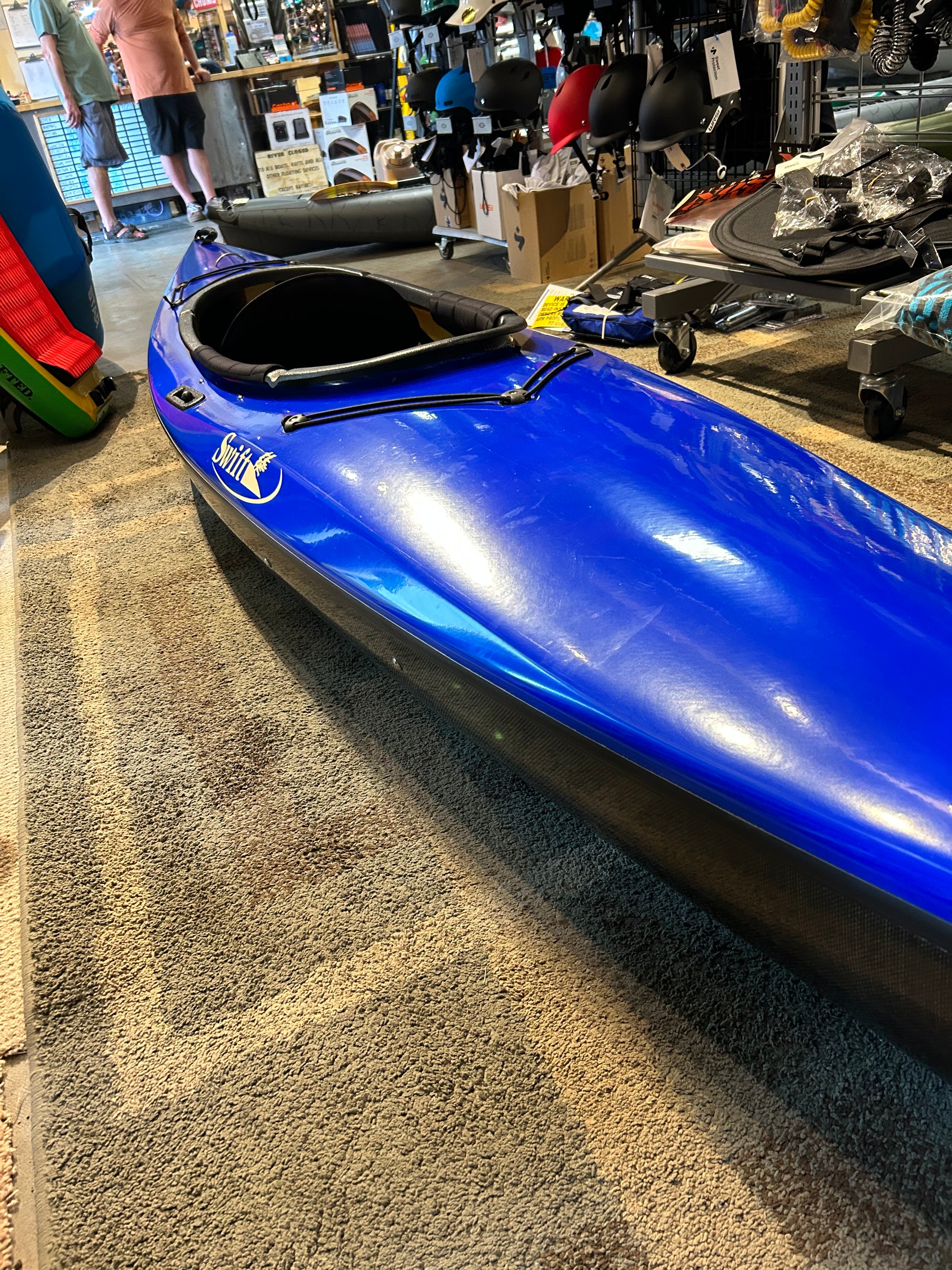 A lightweight Consignment Swift Adirondack 12 LT kayak by 4CRS is displayed indoors on a gray carpet in a store, with various other outdoor equipment and helmets visible in the background. The sleek blue kayak features a narrower cockpit and an advanced deck design for enhanced performance.