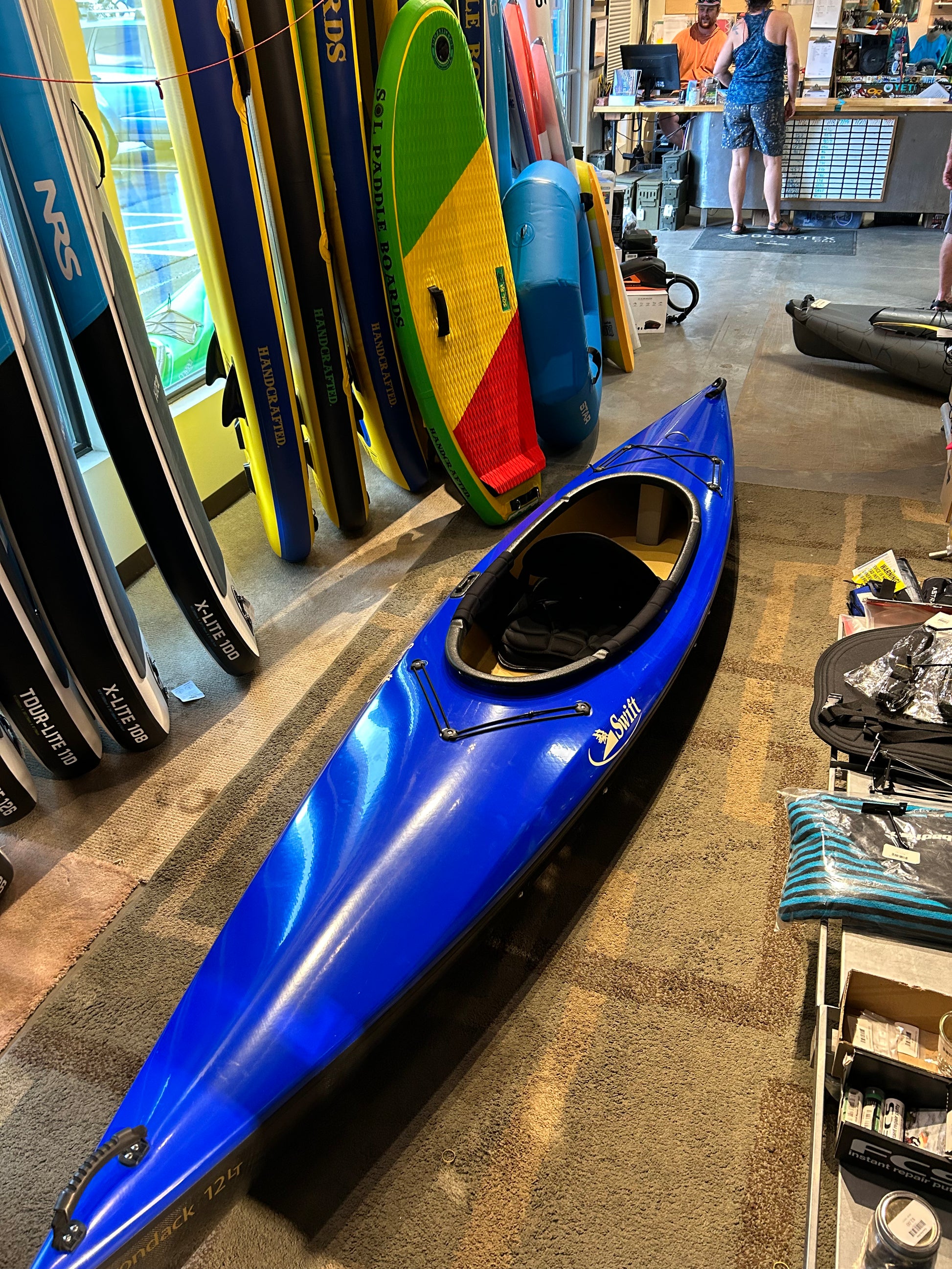 The Consignment Swift Adirondack 12 LT by 4CRS, featuring a sleeker deck design in blue, is displayed on the floor of the store, surrounded by paddleboards and other water sports equipment.