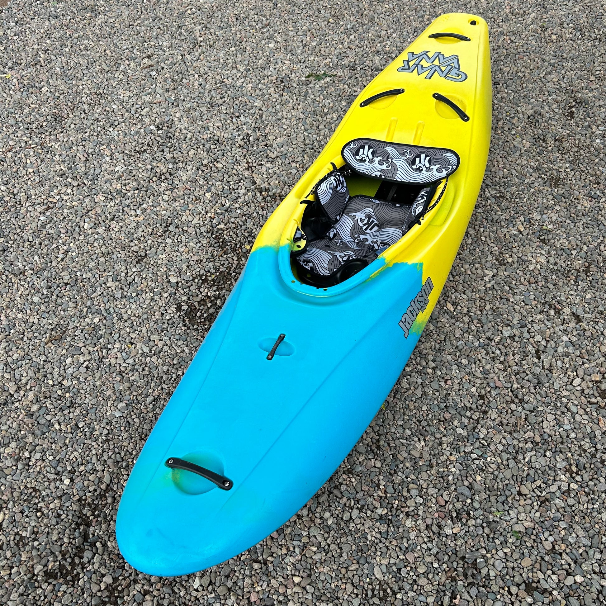 A yellow and blue Demo Gnarvana SM kayak from Jackson Kayak's 2024 used demo fleet, featuring black-and-white patterned seating, is placed on a gravel surface.