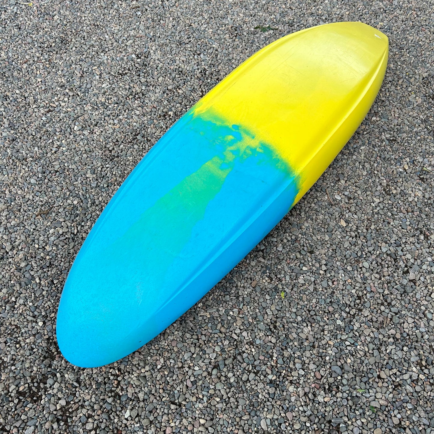 A Jackson Kayak Demo Gnarvana SM, featuring a yellow and blue color gradient reminiscent of the 2024 demo fleet boats, lying on a gravel surface.