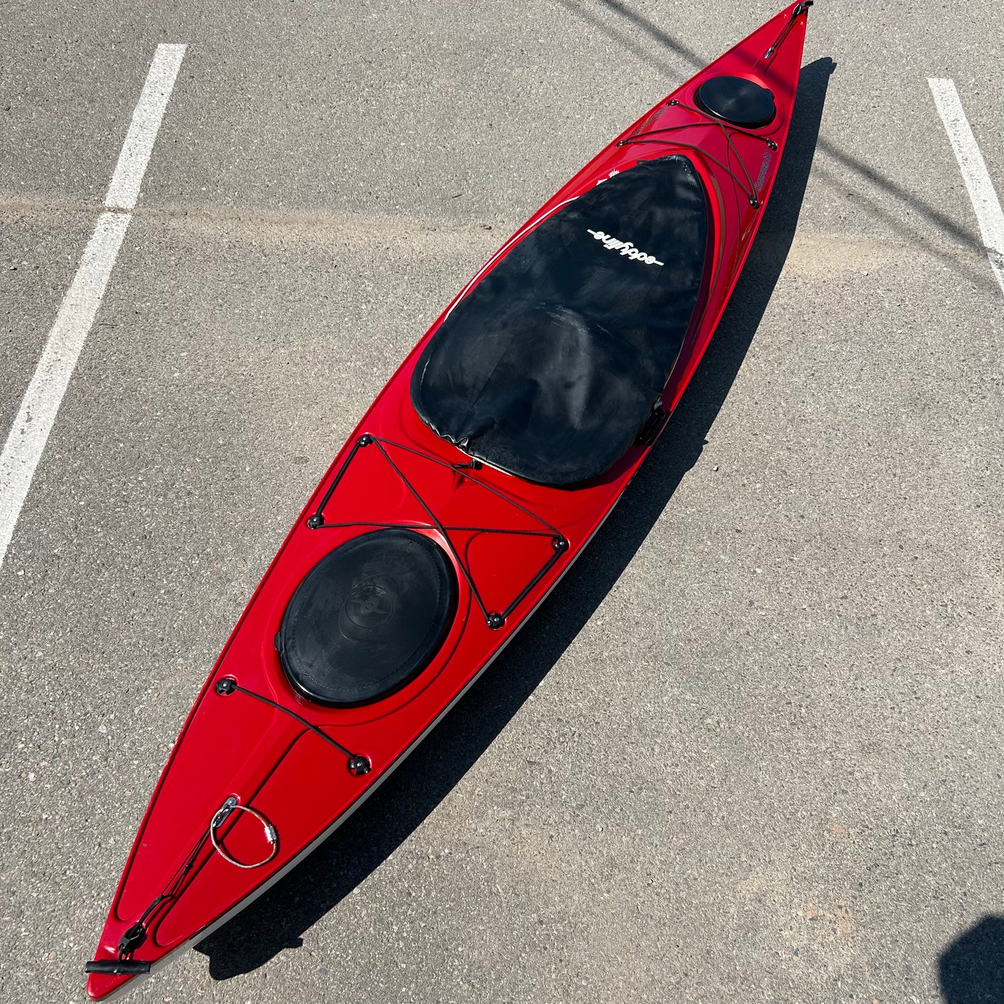 A Consignment Eddyline Sandpiper 130 with black compartment covers is placed on a concrete surface, marked with parking lines.