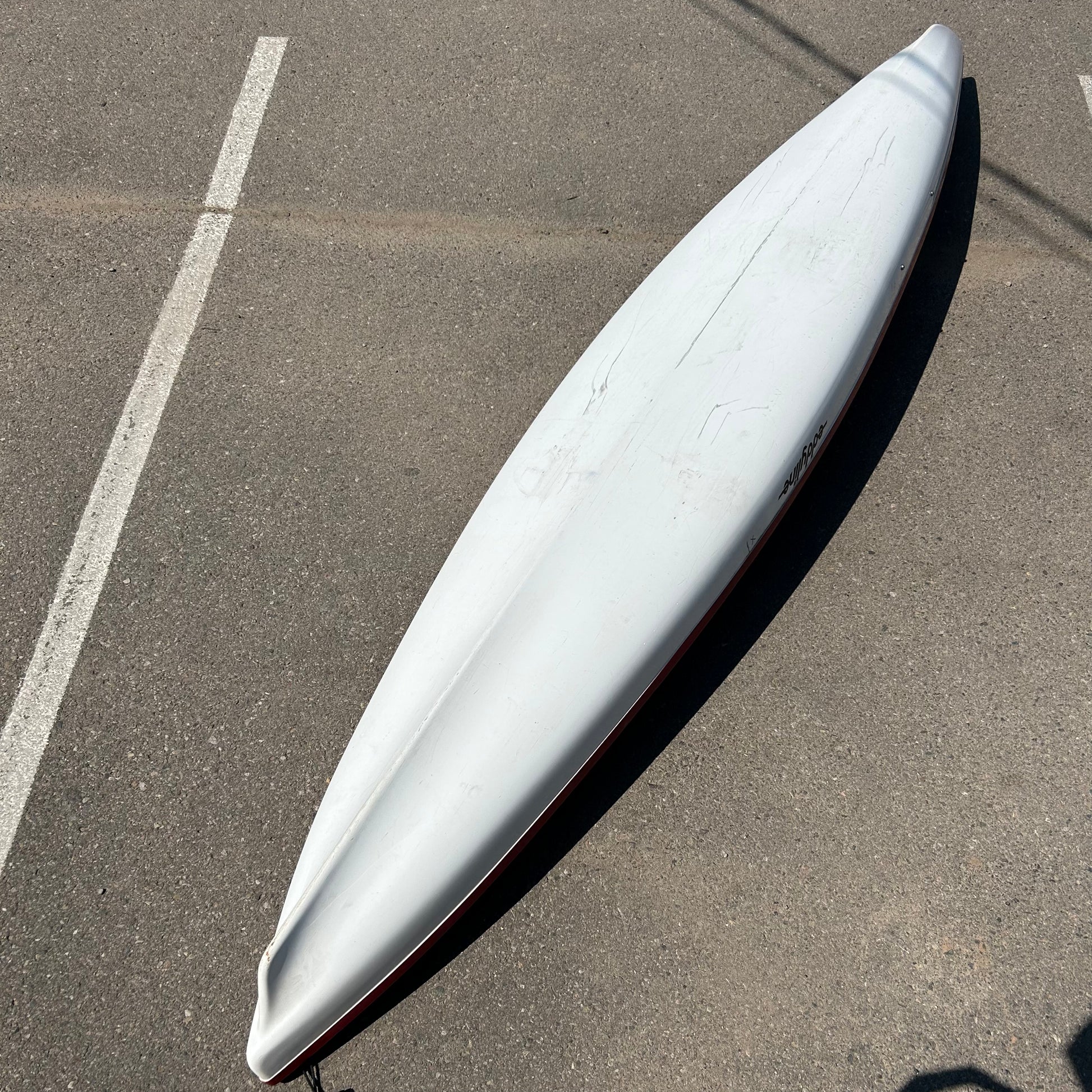 A long white surfboard lies on its underside in an empty parking lot, showing visible wear and scratches. Nearby, a Consignment Eddyline Sandpiper 130 fishing kayak by Eddyline awaits adventures on lakes and rivers.