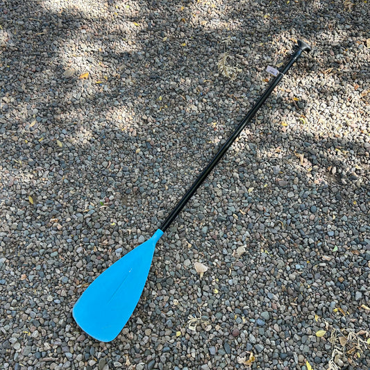 A Demo Glide SUP Paddle by Badfish, featuring a blue and black design, in decent shape, lies on a gravel outdoor surface with scattered small leaves and shadows.