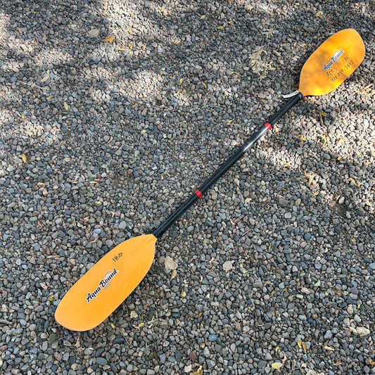 A yellow AquaBound Demo Shred kayak paddle with a Carbon Fiber Shaft and Fiberglass Reinforced Nylon Blade lies on a gravel surface in dappled sunlight.