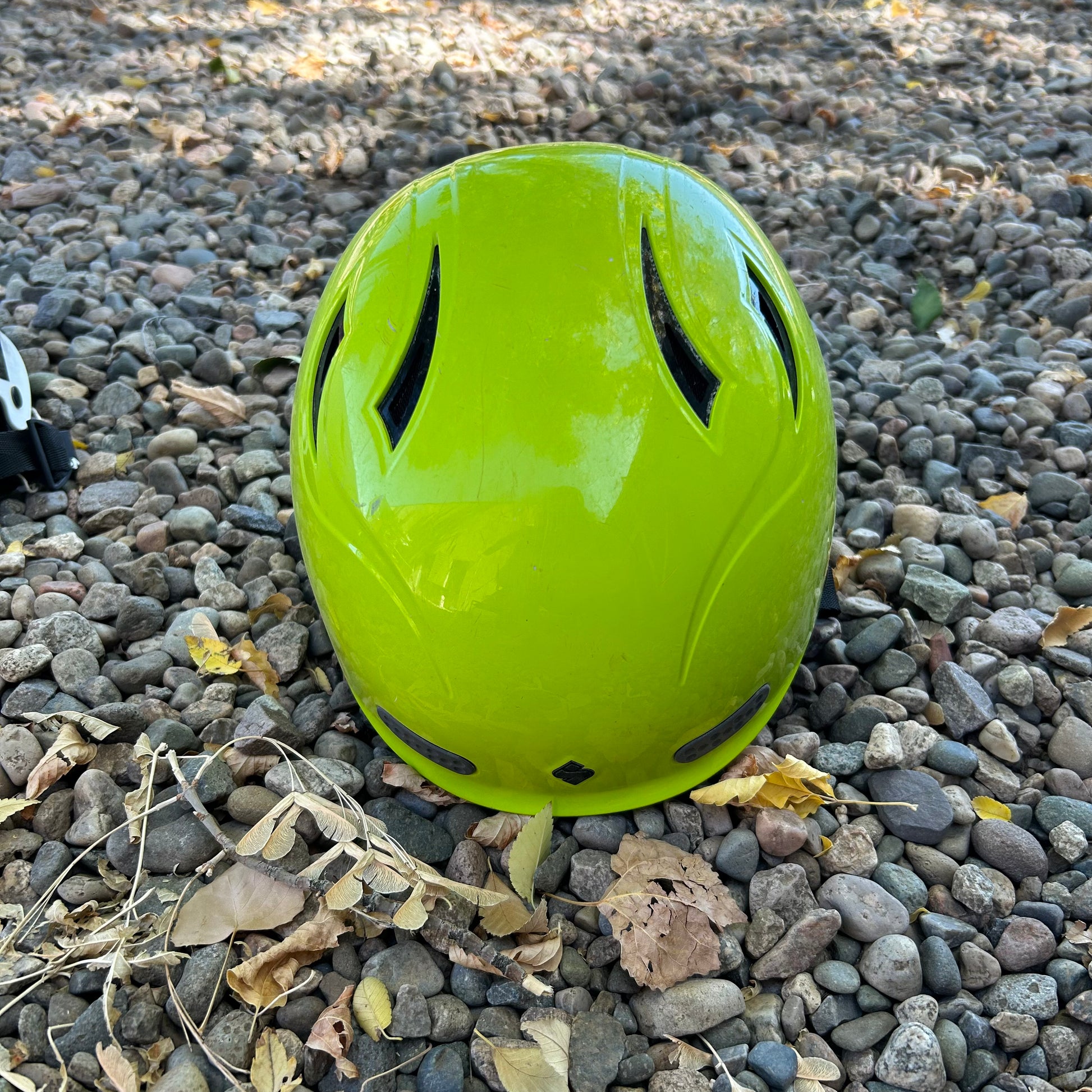 A bright green Demo Wanderer Helmet from Sweet with an ABS Shell lies on a bed of gravel and scattered leaves under dappled sunlight.