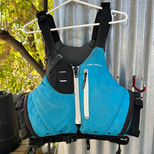 A blue Demo Ceiba PFD by Astral, featuring black straps and a white zipper, is hung on a white clothes hanger against a corrugated metal background, available for local pickup.