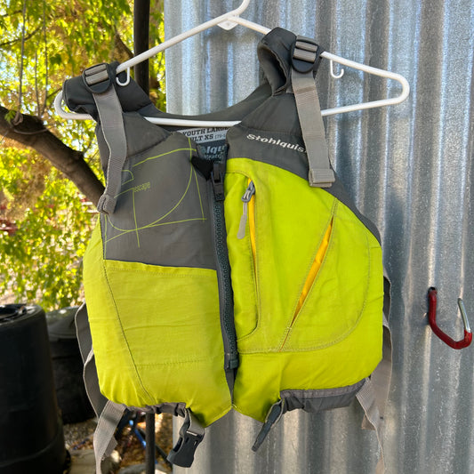 A vibrant green and gray Stohlquist Demo Escape Youth PFD hangs on a white hanger. It features a front zipper and several pockets, set against a background of a metal corrugated wall and trees.