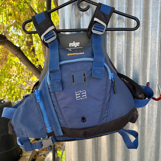 A blue and black life jacket hangs on a hanger. This Demo Edge PFD by Stohlquist features multiple straps, pockets, and mesh panels. Previously part of last year's demo equipment fleet, it remains in excellent condition, showcasing the quality craftsmanship associated with the brand name "Stohlquist".