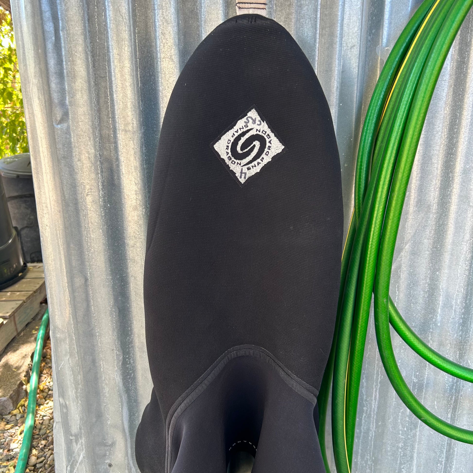 A Snap Dragon Demo River Trek Skirt SM Tube with a white diamond-shaped logo hangs on a metal wall next to a coiled green garden hose, ready for the next River Trek adventure.