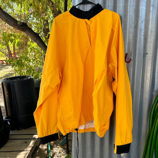 The NRS Demo Youth Splash Jacket, featuring a vibrant yellow design with black accents and a hanging tag, is displayed on a corrugated metal wall. In the background, lush green trees and a black container can be seen, evoking the feel of equipment used at a kayak school.