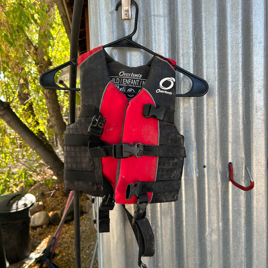 The Stohlquist Demo Overtons Child 33-55lbs life jacket, in red and black, from last year's demo fleet is hanging on a black hanger against a corrugated metal background. A tree and some outdoor items are partially visible in the background, offering a genuine glimpse for local-pickup customers.