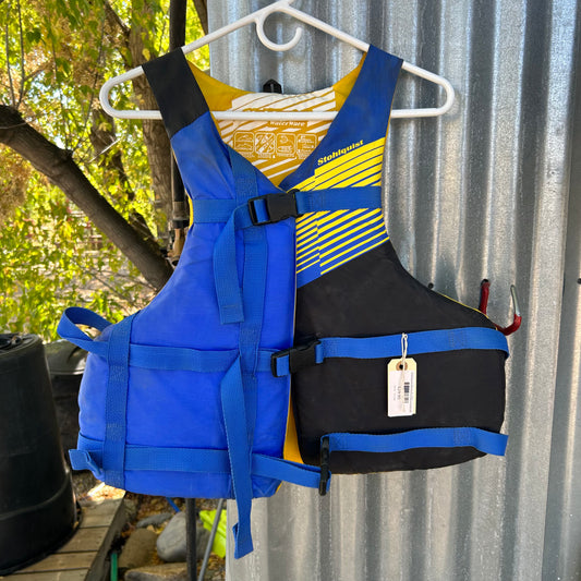 A Stohlquist Demo Fit PFD Oversize, in blue and black with blue straps, hangs on a white hanger against a corrugated metal wall.