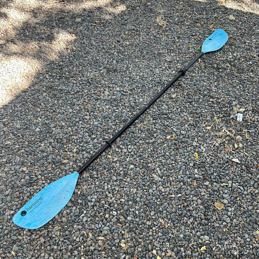 The Harmony Demo Sea Passage Paddle, featuring blue blades and a black shaft, is in good condition and lies on a gravel surface.