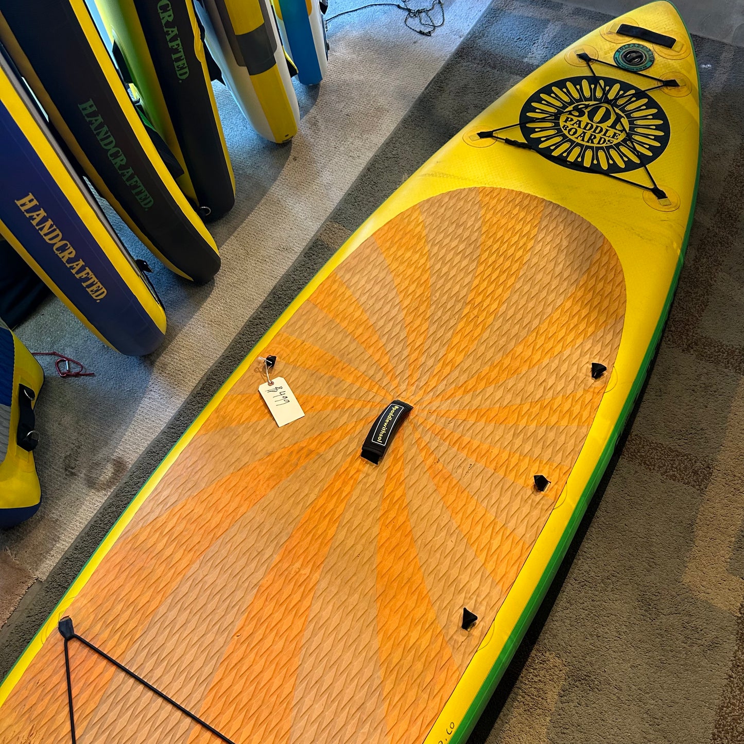 The SOL Paddle Boards Demo Sol Train Classic, a yellow and orange stand-up paddleboard with a sunburst pattern, is displayed upright among other boards in a store and priced at $999.