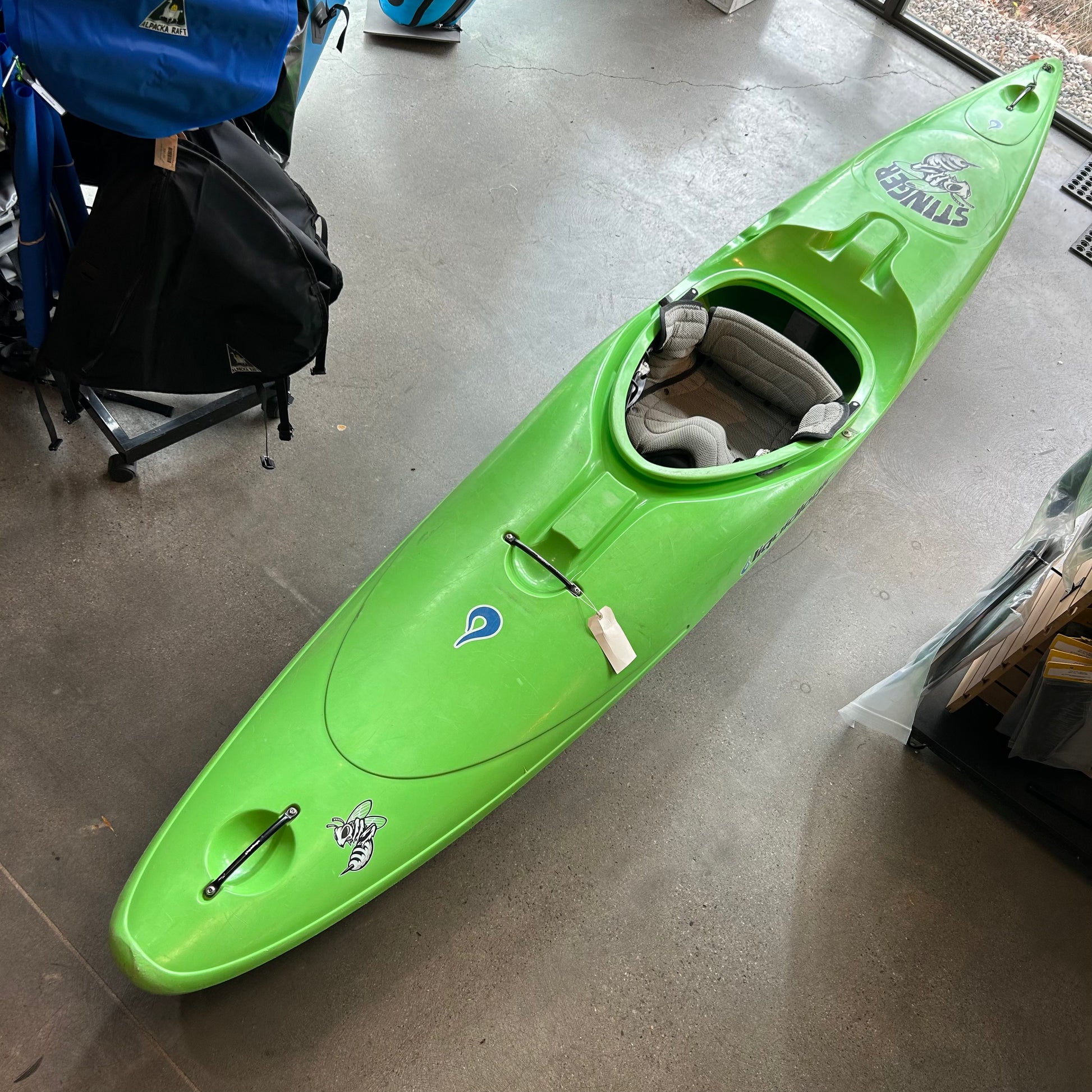 A green LiquidLogic Used Stinger kayak with stickers and a price tag is on the store floor, surrounded by backpacks. Part of our consignment collection, it's perfect for local pickup.