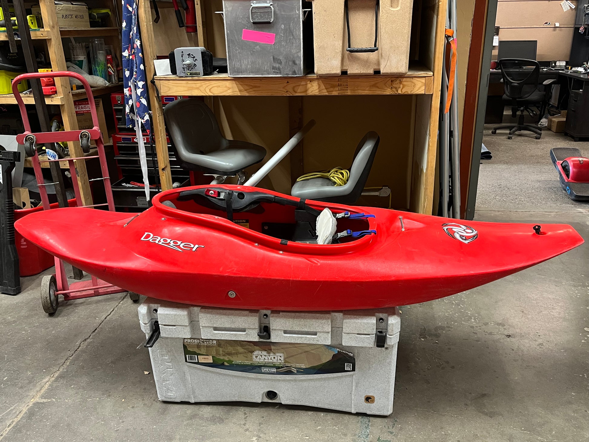 A Consignment Dagger ID 7.0 kayak in red sits on a white cooler in a garage, surrounded by various tools and equipment.