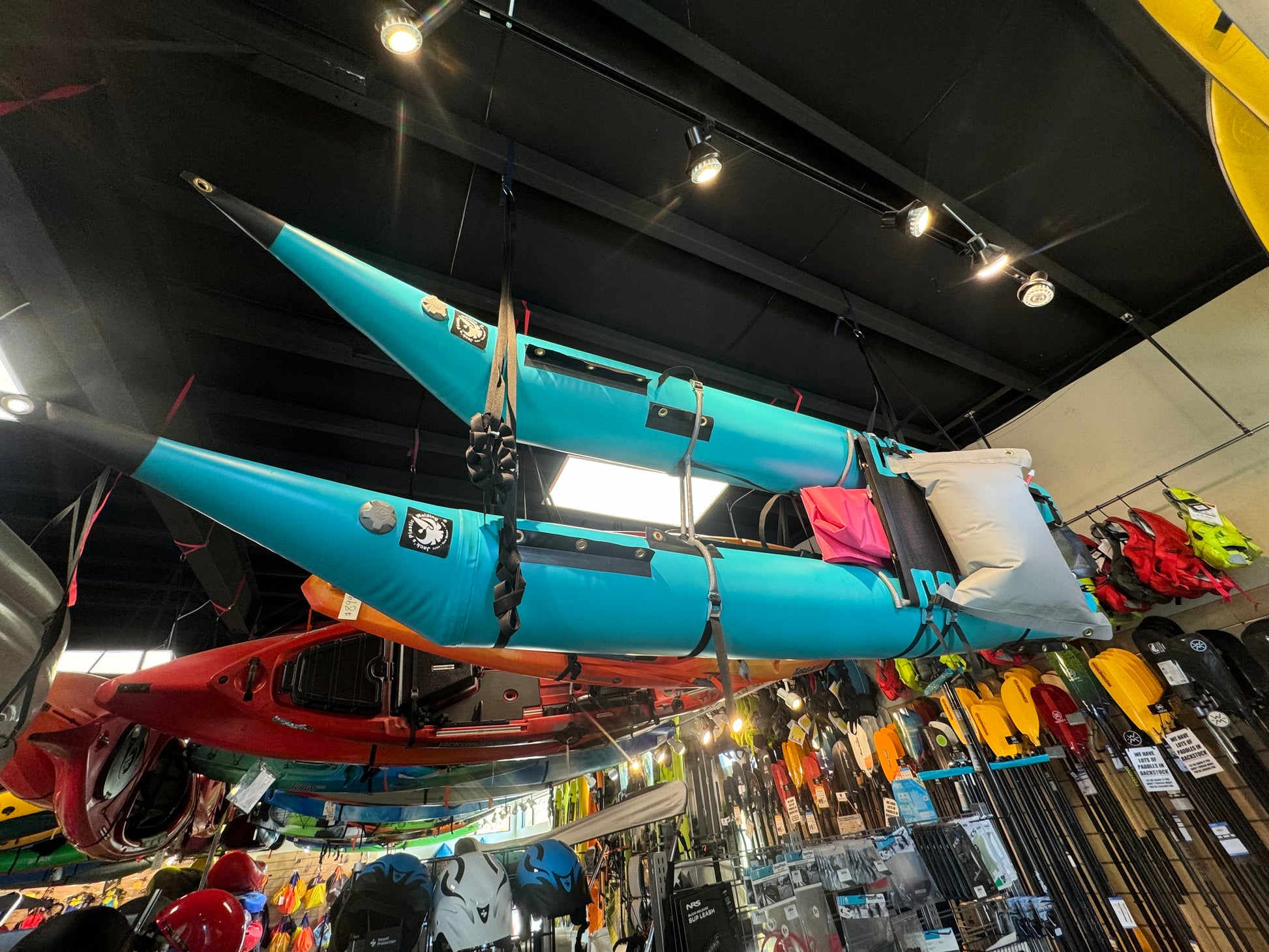 A blue Jacks Plastic Welding "Pack Cat" inflatable kayak in cataraft style hangs from a store ceiling, surrounded by outdoor gear displayed on walls and shelves.