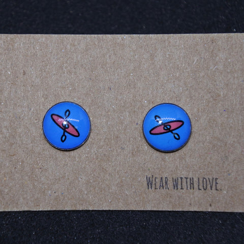 A pair of Kayak Earring Studs by Kingdom Outdoor Products, featuring a round design with a blue background and a red spinning top motif, presented on a brown card bearing the words "Wear with love.