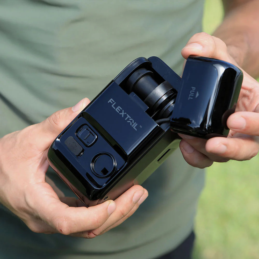 A person holds a black MAX Electric Boat Pump from Flextail with an open compartment, revealing internal components. One hand is removing a detachable cover labeled "PULL." The device, known for its compact design and high-speed motor, is set against a green grassy background.