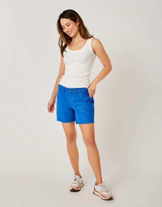 Wearing Carve's Oahu 4" Short and a white tank top, a person stands casually with hands in pockets, embodying the perfect vacation vibe with laid-back sneakers.