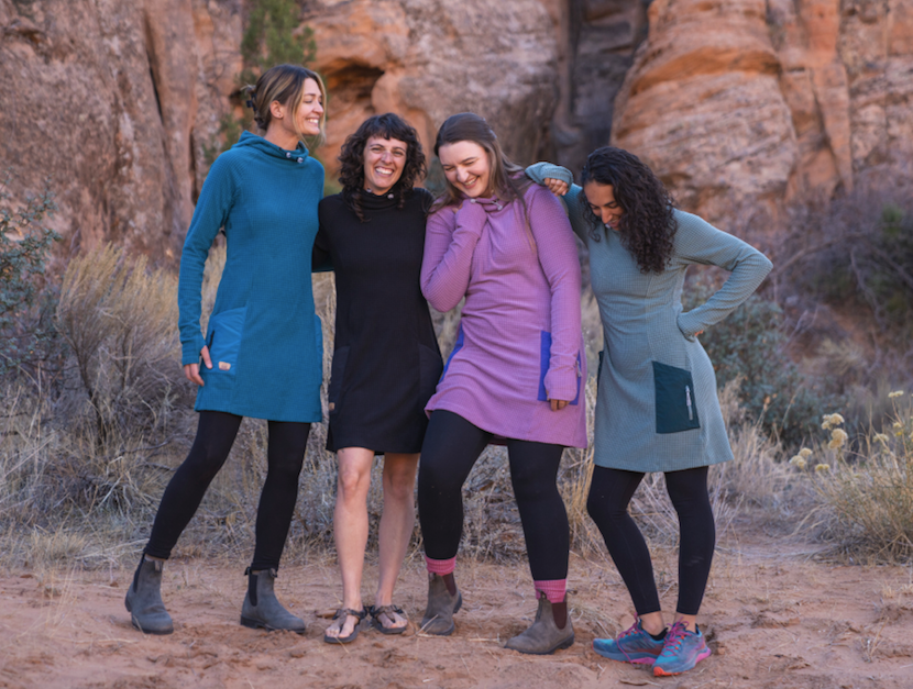 Four individuals wearing Desert Wild's Moab Trail Dress stand and smile on a sandy terrain with rocky cliffs in the background.